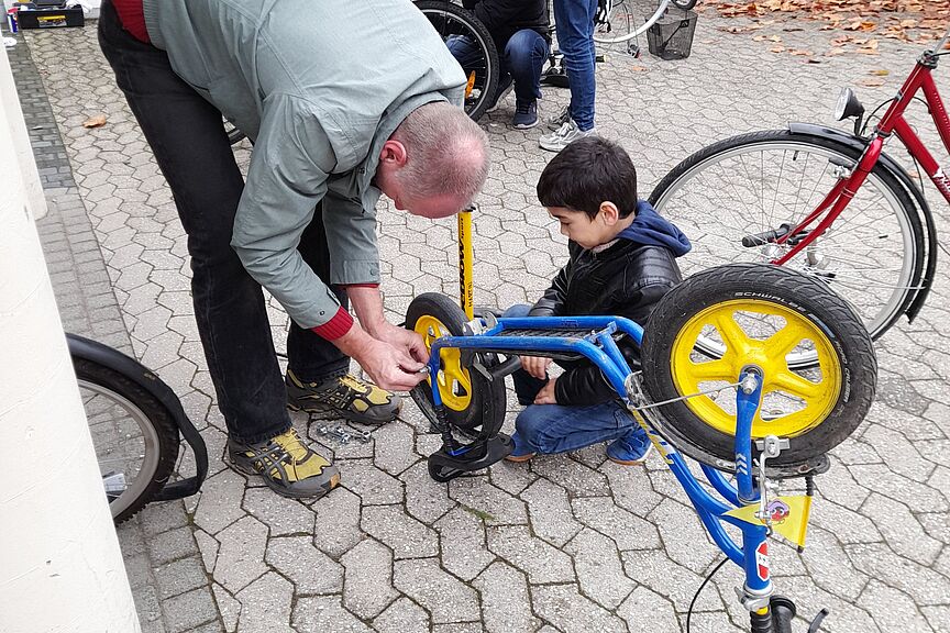 Fahrradausgabe Samstagstreff Das Bild zeigt einen Mann und einen kleinen Jungen, der Mann repariert das kleine Fahhrad des Jungen, der gespannt daruaf wartet