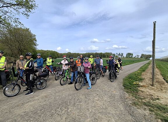Das Bild zeigt eine Gruppe von 20 Radfahrenden der Hofladentour beim Stopp vor einer blüghenden Apfelplantage am Ortsrand von Werthhoven