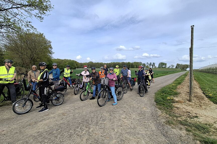 Hofladentour Das Bild zeigt eine Gruppe von 20 Radfahrenden der Hofladentour beim Stopp vor einer blüghenden Apfelplantage am Ortsrand von Werthhoven