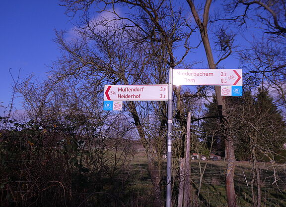 Das Bild zeigt einen roten Radwegweiser, wie er in NRW üblich ist, der in zwei Richtungen nach Heiderhof bzw. Ließem zeigt. Daran befestigt das Logo des NGL: schematischer Wachtturm in Kreis mit stilisiertem Rheinverlauf auf blauem Grund.