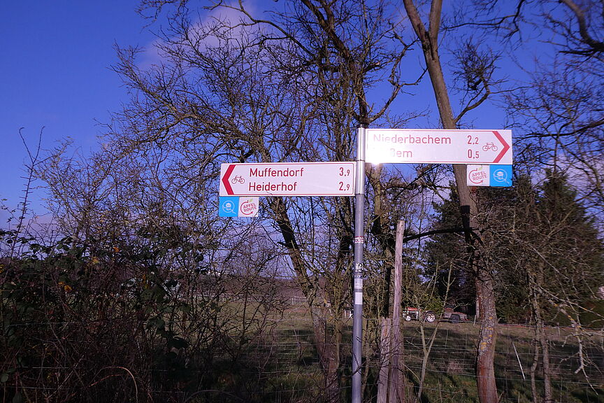 Wegweise NGL-Radweg Das Bild zeigt einen roten Radwegweiser, wie er in NRW üblich ist, der in zwei Richtungen nach Heiderhof bzw. Ließem zeigt. Daran befestigt das Logo des NGL: schematischer Wachtturm in Kreis mit stilisiertem Rheinverlauf auf blauem Grund.