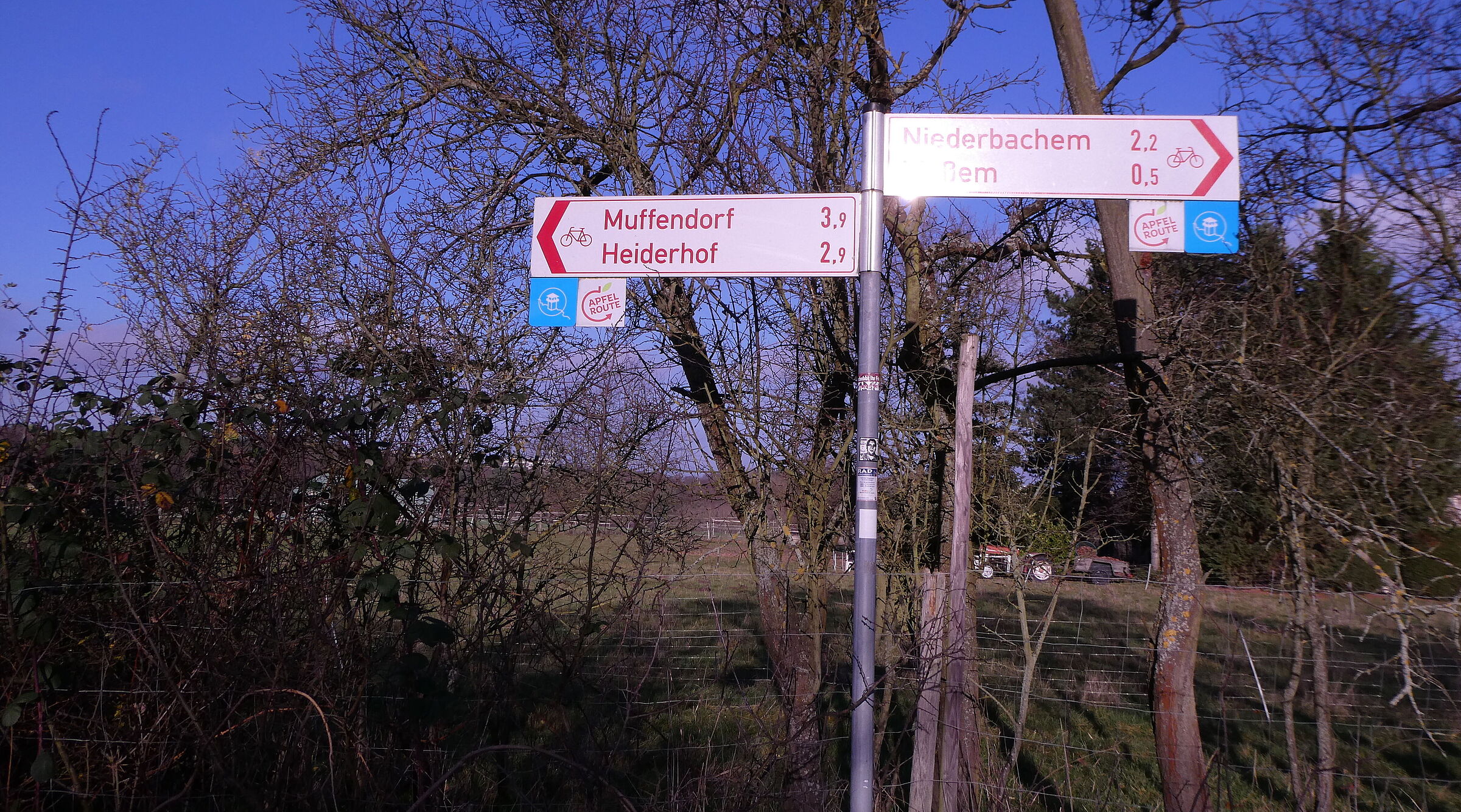 Das Bild zeigt einen roten Radwegweiser, wie er in NRW üblich ist, der in zwei Richtungen nach Heiderhof bzw. Ließem zeigt. Daran befestigt das Logo des NGL: schematischer Wachtturm in Kreis mit stilisiertem Rheinverlauf auf blauem Grund.