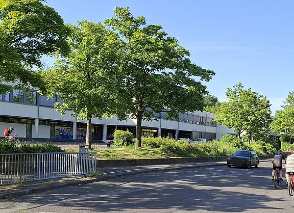 Das Bild zeigt die Straße vor der Schule in Berkum, mit einigen Radfahrern im Vordergrund und dem Schulgebäude im Hintergrund