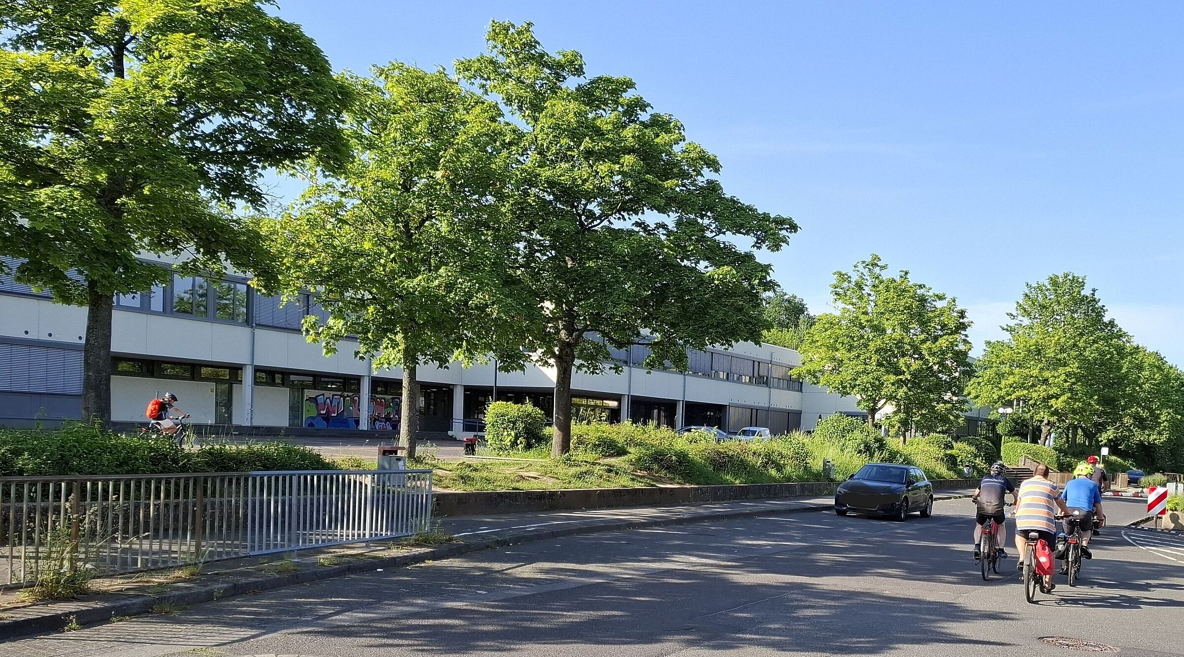 Das Bild zeigt die Straße vor der Schule in Berkum, mit einigen Radfahrern im Vordergrund und dem Schulgebäude im Hintergrund