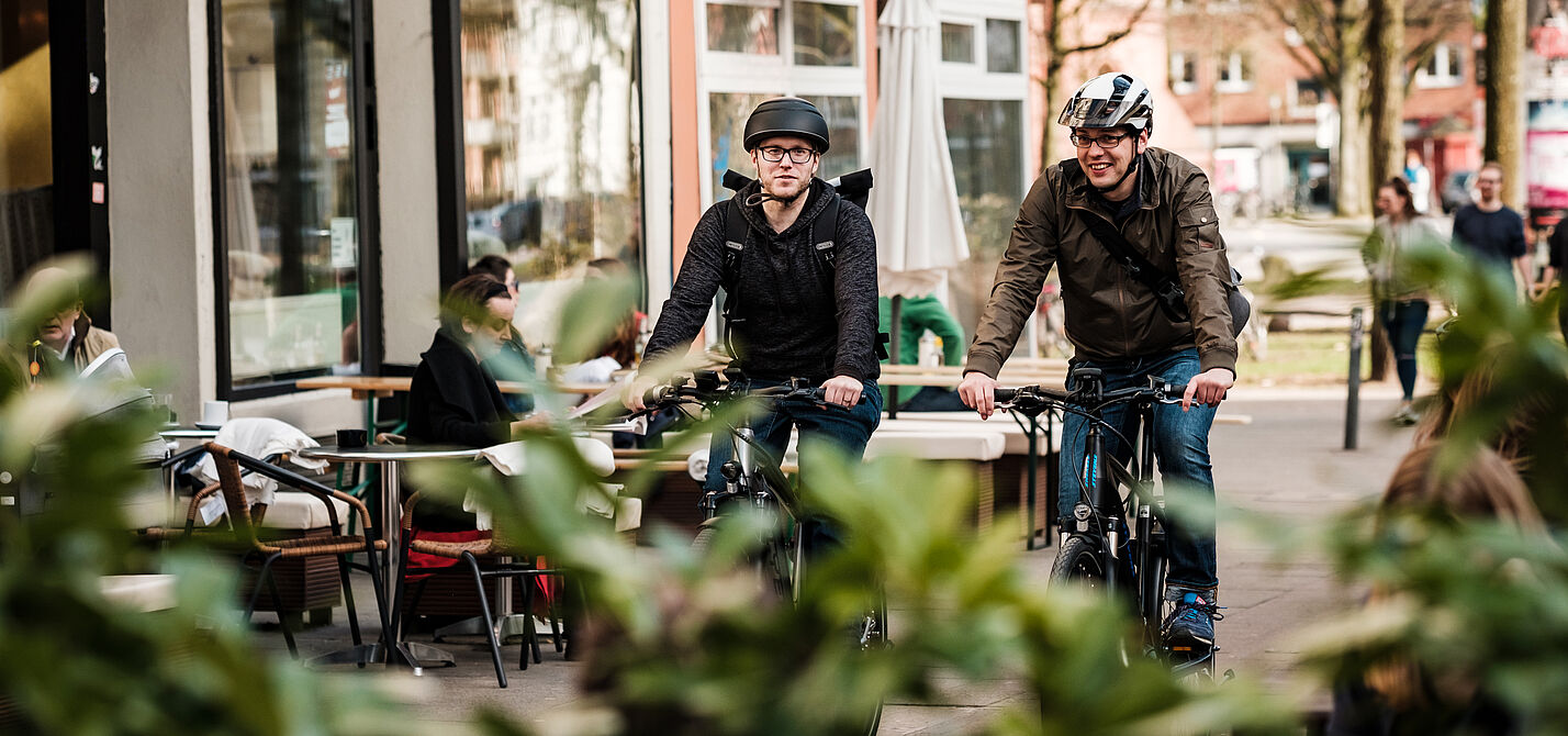 Zwei Männer mit Helm fahren auf einem Radweg im urbanen Raum
